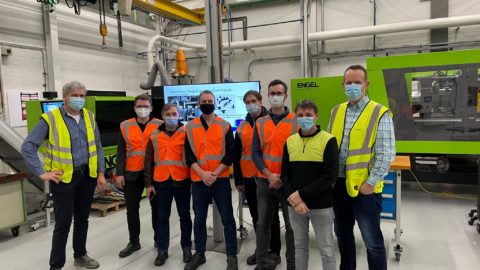 Two groups of men stand together in a factory setting , five are wearing high vis orange vests, three with high vis neon. All are wearing masks on their faces.